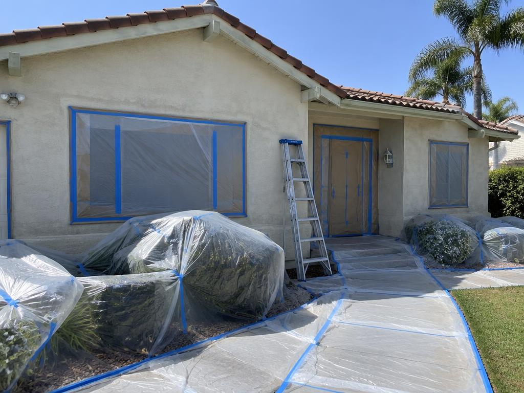 House exterior prepared for painting, with windows and door masked and shrubs covered in plastic sheeting